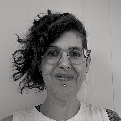 Black and white portrait of a smiling person with curly hair, glasses, and facial piercings, standing in front of a white paneled wall.